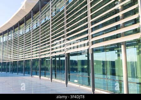 Gekrümmte Außen-Glasfassade eines modernen Firmengebäudes mit horizontalen Metall-Sonnenlamellen und Stahlstützsäulen, die das Sonnenlicht reflektieren. Stockfoto