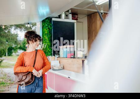 Stylische lockige brünette Frau, die während eines Stadtspaziergangs an einem Outdoor-Kiosk Dessert auswählt Stockfoto