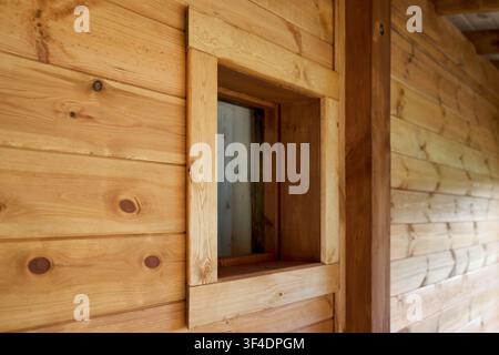 Nahaufnahme eines kleinen quadratischen Glasfensters, das in eine rustikale Holzwand aus gezapften Kiefernholzplanken mit heller Naturlackierung integriert ist, das ein w Stockfoto