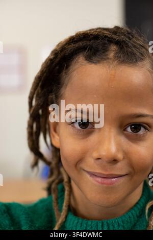 Afroamerikanisches Kind lächelt in die Kamera im Klassenzimmer mit verschwommenem Poster, in grünem Pullover Stockfoto