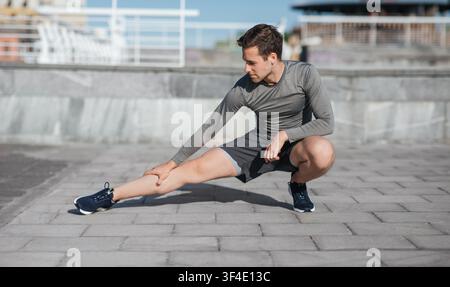 Sportlicher Körper und gesunder Lebensstil. Mann hockt und tut Beinübungen auf der Straße Stockfoto