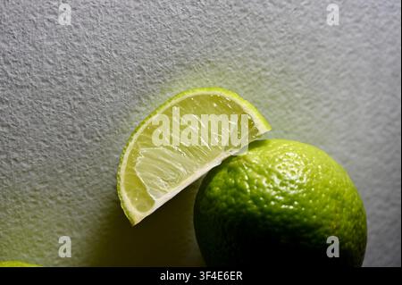 Eine Makro-Nahaufnahme eines saftigen Limettenwürfels, der auf einer ganzen frischen grünen Limette ruht, vor einem neutralen strukturierten Hintergrund, betont durch dramatische Richtungsbeleuchtung Stockfoto