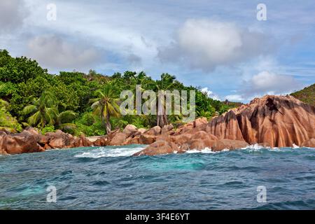 Tropische Küste der Insel Curieuse, Seychellen, wo türkisfarbene Wellen an geschwungenen Granitfelsen brechen, unter Kokospalmen und üppigem Wald. Stockfoto