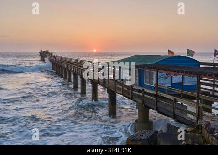 Ein glühender goldener Sonnenuntergang umrahmt den historischen Holzsteg in Swakopmund, Namibia, während mächtige Wellen des Atlantischen Ozeans gegen seine Pfosten schlagen. Stockfoto