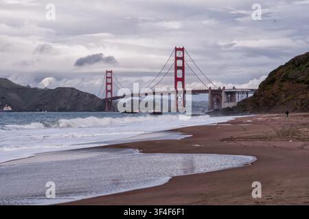 Die goldene Brücke steht kräftig vor dem bewölkten Himmel. Wellen brechen mit weißem Schaum an der sandigen Küste. Ferne Hügel rahmen die ikonische Spannweite. Die Stimmung ist ruhig, still, sl Stockfoto