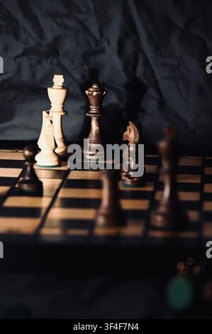 Ein stimmungsvolles, gedämpftes Foto eines Holzschachbretts mit Fokus auf die Hauptfiguren, einschließlich des weißen Königs, des Springers und der schwarzen Dame. Tiefe Schatten, atmosphärische dramatische Beleuchtung und ein dunkler, zerknitterter Hintergrund erzeugen ein starkes Gefühl von Strategie, Wettbewerb, Spannung und Intellekt. Perfekt für Konzepte, die sich auf Geschäftsstrategie, Entscheidungsfindung, Taktik und klassische Tischspiele beziehen. Stockfoto