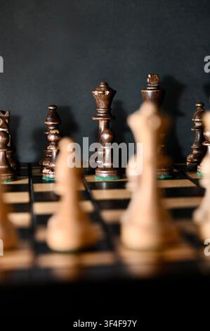 Eine Nahaufnahme, die die schwarzen Holzschachfiguren mit scharfer Fokussierung auf König, Dame und Läufer einfängt, während die gegnerischen weißen Figuren im Hintergrund weich unscharf bleiben. Atmosphärische Low-Key-Beleuchtung hebt die feine Holztextur hervor und erzeugt ein dramatisches Gefühl von Konfrontation, Strategie, Führung und Weitsicht. Perfekte visuelle Darstellung für Geschäftsrivalität, taktische Planung und wettbewerbsfähigen Intellekt. Stockfoto