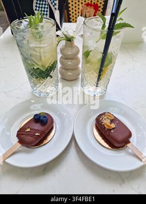 Zwei elegante Schokoladen-Popcake stehen auf dem Tisch in der Nähe hoher Gläser mit erfrischender Minz-Mojito-Limonade mit Eis. Das Dessert ist mit Erdnuss garniert Stockfoto