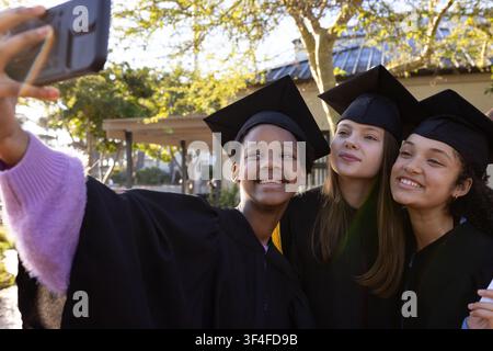 Verschiedene Teenager-Schülerinnen machen ein Selfie auf dem Campus mit Smartphone, Mützen, Roben, Quasten Stockfoto