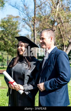 Zufriedener Elternteil feiert den Abschluss des Studiums mit einem stolzen Absolventen, der Hut und Talar trägt, im Freien Stockfoto