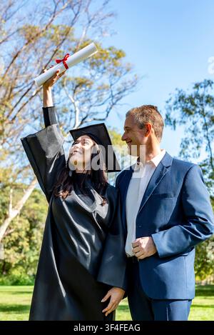 Vertikales Porträt einer Studentin in Hut und Robe, die ihr Diplom neben einem stolzen Mann mittleren Alters hochhält Stockfoto