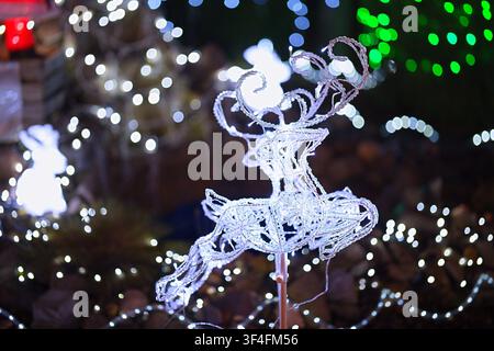 Eine leuchtende weiße Weihnachtsfigur, die ein Rentier darstellt, in einer nächtlichen Außeneinrichtung, mit Engelsfigur, Lichterkette, aus einer Frontalperspektive gesehen, Stockfoto