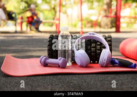 Fitnessgerät für draußen mit violetten Hanteln, Wasserflasche und Kopfhörern auf einer roten Yogamatte im Park Stockfoto