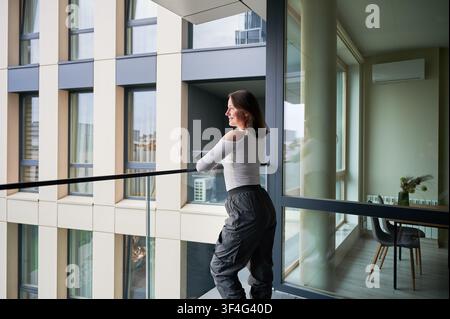 Junge Frau steht auf dem Apartmentbalkon und hält ein Smartphone. Frau betrachtet die Stadtansicht nahe einem offenen Fenster, genießt einen ruhigen Moment, urbaner Lebensstil, modernes Wohnkonzept Stockfoto