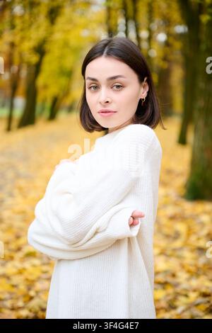 Porträt einer jungen Frau, die in einen weichen weißen Pullover gehüllt ist und in einem Herbstpark mit gelben Blättern und verschwommenen Bäumen im Hintergrund posiert. Perfekt für Mode, gemütliches Lifestyle, Schönheit, saisonale Werbung und Anzeigen Stockfoto