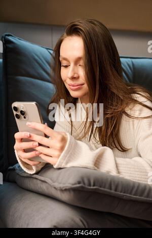 Junge Frau entspannt sich auf dem Sofa und verwendet ein Smartphone, lächelt beim Surfen oder Schreiben von Nachrichten, repräsentiert modernen Lebensstil, digitale Kommunikation, Freizeit und gemütlichen Innenkomfort Stockfoto