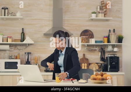 Freiberufler eilt vor der Arbeit durch das Frühstück, isst schnell und überprüft die Zeit, was eine hektische Morgenroutine vor der Arbeit zeigt. Stockfoto