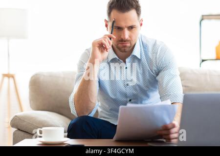 Nachdenklicher junger Mann, der am Schreibtisch sitzt und Dokumente ansieht Stockfoto