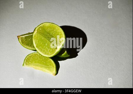 Eine hochkontrastierte Makrofotografie mit frischen grünen Limettenwürfeln auf einem weißen strukturierten Hintergrund. Helles, gerichtetes Sonnenlicht beleuchtet das saftige Zitrusmark und erzeugt scharfe, dunkle geometrische Schatten auf der Oberfläche. Eine moderne, minimalistische Komposition, perfekt für Food-Fotografie, Sommergetränkerezepte, Cocktailkonzepte, Barmenus und frische Zitrusmarken. Stockfoto
