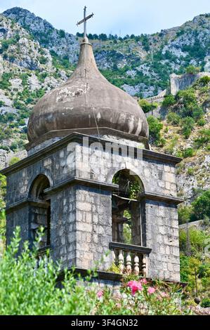 Vertikale Aufnahme eines alten steinernen christlichen Kirchenglockenturms mit einer Kreuzkuppel vor felsigen Bergen in Kotor, Montenegro. Ideal für Reisethemen, Geschichte, Religion und Balkanarchitektur. Stockfoto