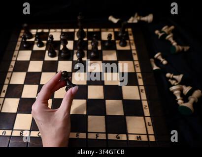 Eine obere, stimmungsvolle Ansicht der Hand eines Spielers, die eine schwarze Schachfigur hält und eine Entscheidung auf einem hölzernen Schachbrett trifft. Dunkle und helle Schachfiguren sind in einer taktischen Position angeordnet, wobei eroberte Figuren neben dem Brett ruhen, was Geschäftsplanung, Führung, Entscheidungsfindung und Strategie symbolisiert. Stockfoto