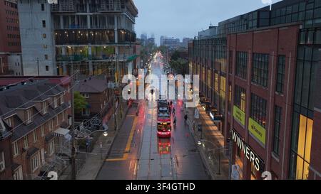 TORONTO, KANADA – 10. MAI 2024: Luftperspektive, die eine nasse Stadtstraße im Regen hinunterblickt, mit einer roten Straßenbahn, die entlang der Schienen an Einzelhandelsgeschäften vorbeifährt Stockfoto