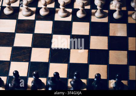 Hochwinklige Flachlay-Ansicht eines Holzschachbretts mit allen Figuren in Startposition angeordnet. Der dramatische Kontrast zwischen hellen und dunklen Tönen mit subtilen Schatten symbolisiert strategische Planung, Geschäftskonkurrenz, taktisches Denken, Entscheidungsfindung und intellektuelle Herausforderung. Stockfoto