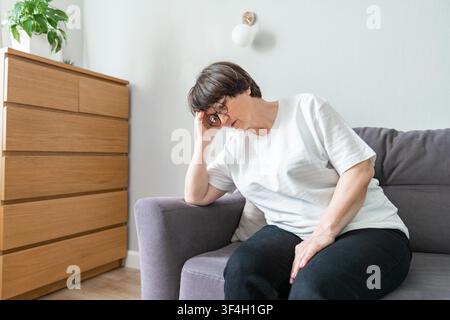 Müde, deprimierte ältere Frau sitzt auf der Couch im Wohnzimmer und fühlt sich verletzt und einsam. Ältere, weißhaarige Oma berührt die Stirn und leidet unter starken Kopfschmerzen oder erinnert sich an schlechte Erinnerungen Stockfoto