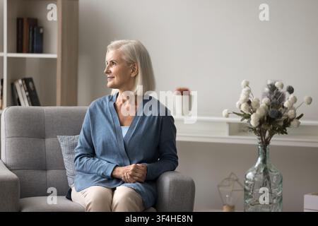 Verträumte reife weibliche Rentnerin entspannt sich im Sessel im Wohnzimmer Stockfoto