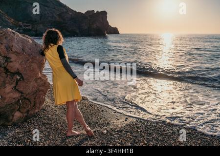 Frau am Strand bei Sonnenuntergang mit Blick auf den Ozean, während sie sich am Abend auf einen Felsen lehnt Stockfoto