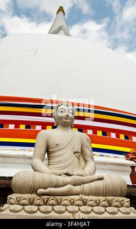 Sitzende Stein-Buddha-Statue vor einer großen weißen buddhistischen Stupa, die in einer farbenfrohen buddhistischen Flaggenbanner gewickelt ist, Sri Lanka. Stockfoto