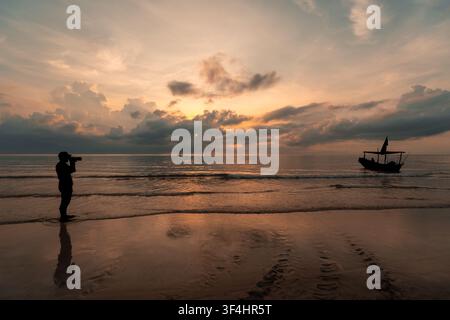 Ein einsamer Fotograf in Silhouette fängt den atemberaubenden goldenen Sonnenaufgang über einem tropischen Strand ein, mit einem traditionellen Fischerboot, das sanft auf der C Stockfoto