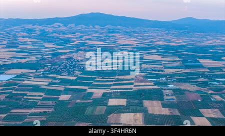 Ein erhöhtes Panorama zeigt bewirtschaftete Felder in geometrischen Blöcken, eine kleine Siedlung und geschichtete Bergkämme unter einem weichen pastellfarbenen Abendhimmel. Stockfoto