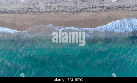 Eine Blick-von-oben-Küstenszene zeigt schaumige, türkisfarbene Wellen, die über flaches Wasser auf einen leeren Streifen hellen Sandes in Nea Moudania, Griechenland, zurollen. Stockfoto