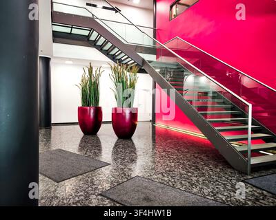 Moderne Bürolobby mit roter Akzentwand und Glastreppe, zeitgenössisches Firmeninterieur mit Granitboden, zylindrischen Säulen und großer Pflanze Stockfoto