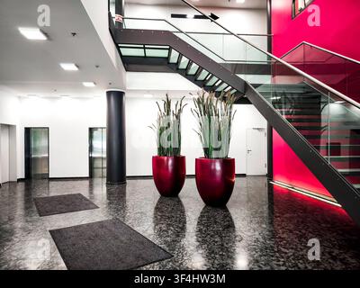 Moderne Bürolobby mit roter Akzentwand und Glastreppe, zeitgenössisches Firmeninterieur mit Granitboden, zylindrischen Säulen und großer Pflanze Stockfoto