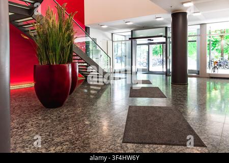 Moderne Bürolobby mit roter Akzentwand und Glastreppe, zeitgenössisches Firmeninterieur mit Granitboden, zylindrischen Säulen und großer Pflanze Stockfoto