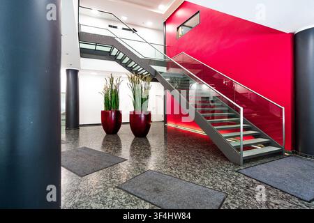 Moderne Bürolobby mit roter Akzentwand und Glastreppe, zeitgenössisches Firmeninterieur mit Granitboden, zylindrischen Säulen und großer Pflanze Stockfoto