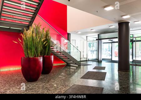 Moderne Bürolobby mit roter Akzentwand und Glastreppe, zeitgenössisches Firmeninterieur mit Granitboden, zylindrischen Säulen und großer Pflanze Stockfoto