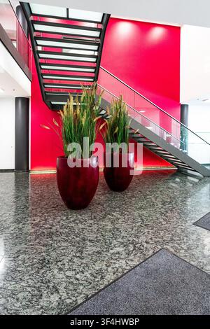 Moderne Bürolobby mit roter Akzentwand und Glastreppe, zeitgenössisches Firmeninterieur mit Granitboden, zylindrischen Säulen und großer Pflanze Stockfoto