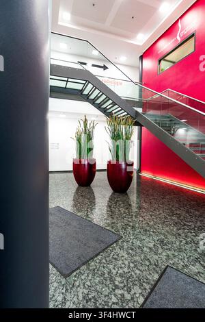 Moderne Bürolobby mit roter Akzentwand und Glastreppe, zeitgenössisches Firmeninterieur mit Granitboden, zylindrischen Säulen und großer Pflanze Stockfoto