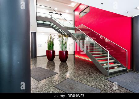 Moderne Bürolobby mit roter Akzentwand und Glastreppe, zeitgenössisches Firmeninterieur mit Granitboden, zylindrischen Säulen und großer Pflanze Stockfoto
