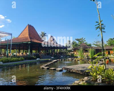 Eine ruhige Außenszene mit traditioneller indonesischer Joglo-Architektur, einem klaren Koiteich mit Stegen und üppiger Vegetation unter einem hellen Bl Stockfoto