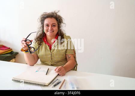 Zufriedene Designerin skizziert und entwickelt Konzepte. Porträt einer lockenköpfigen Künstlerin, die in einem hellen Atelier arbeitet Stockfoto