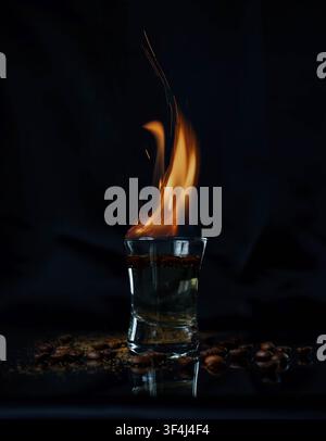 Flammender Schuss von starkem Alkohol mit leuchtend orangefarbenem Feuer, das über einem Glas auf einem schwarzen Hintergrund in dramatischer Low-Key-Beleuchtung brennt. Stockfoto