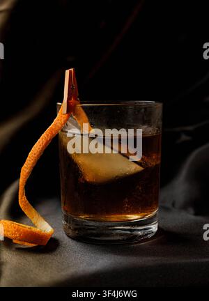 Klassischer Old Fashioned-Cocktail mit Orangenschalengarnitur und großem Eiswürfel, serviert in einem Rocks-Glas vor dunklem Hintergrund. Stockfoto