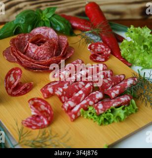 Verschiedene geschnittene Salami und gepfefferte Würste, angeordnet auf einem Holzservierteller mit frischen Kräutern und Gemüse. Traditionelle Charcuterie-Vorspeise. Stockfoto