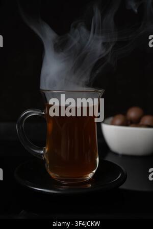 Heißer schwarzer Tee, serviert in einer traditionellen Glastasse mit dramatischem Dampf auf dunklem Hintergrund. Konzept für ein gemütliches warmes Getränk. Stockfoto