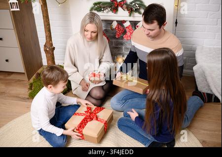 Glückliche Familie tauscht Weihnachtsgeschenke zu Hause aus, Eltern mit Kindern teilen Bastelpapier-Geschenke in der Nähe des geschmückten Kamins, gemütliche Winterurlaubsfeier, Neujahrsüberraschungskonzept. Vigilia Stockfoto