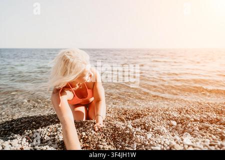 Blonde Strandurlaubs-schöne Frau in Korallen-Bikini, die mit Kieselsteinen an einem sonnigen Ufer neben den blauen Meereswellen spielt Stockfoto